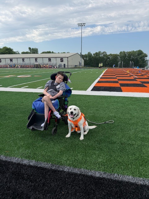Caden in his wheelchair on a football field with Shiloh, his yellow Lab service dog, sitting calmly beside him in an orange service vest